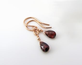 May include: A pair of rose gold drop earrings featuring faceted, teardrop-shaped dark red garnet gemstones. Each earring is suspended from a simple curved ear wire and accented with a small, polished rose gold bead above the gemstone.