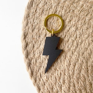 May include: Black lightning bolt keychain with a gold key ring. The keychain is on a woven brown background.
