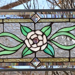 May include: Rectangular stained glass window panel featuring a central white rose with a purple center, surrounded by green leaves and flowing ribbons. The design is framed by clear and textured glass, with diamond-shaped accents at the corners.