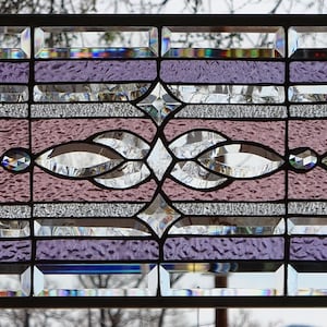 May include: Rectangular stained glass window panel with a black metal frame. The design features a central pattern of lavender and pink glass with clear and textured accents. The panel is approximately 30 inches wide and 12 inches tall.
