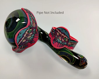 May include: A pink and green glass pipe with a colorful, abstract design. The pipe has a round bowl and a curved stem. The bowl is decorated with a pink and green silicone cover. The stem is decorated with a silicone band with a colorful, abstract design. The text "Pipe Not Included" is visible in the image.