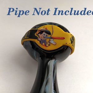May include: A black and blue glass pipe with a yellow band featuring a cartoon image of Pinocchio. The text "Pipe Not Included" is above the pipe. The pipe has a rounded bowl and a long, curved stem.