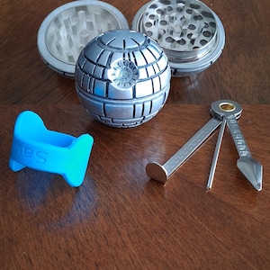 May include: A silver metal herb grinder with a Death Star design, disassembled, next to a blue silicone tool and a metal pipe tool. The grinder is in three pieces.