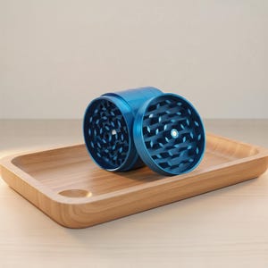 May include: A blue metal grinder with a disassembled top, revealing the interior. The grinder is cylindrical and sits on a wooden tray. The tray has a rounded rectangular shape and a small circular indentation.