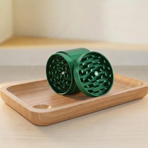 May include: A green herb grinder with a disassembled lid sits on a wooden tray. The grinder is cylindrical with a textured surface and a leaf-shaped pattern on the grinding surface. The tray is rectangular with rounded edges.