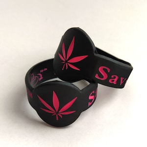 May include: Two black silicone wristbands with a pink cannabis leaf design. One band has the word "Save" in pink. The other band has the words "Flip-On" and a heart symbol in pink.
