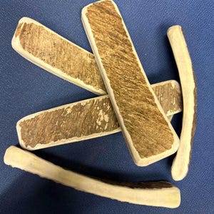 May include: A collection of five natural deer antler chews arranged in a scattered pattern. Each piece features a rough, brown, textured outer surface with smooth, cream-colored edges, showcasing the organic shape and density of the material.