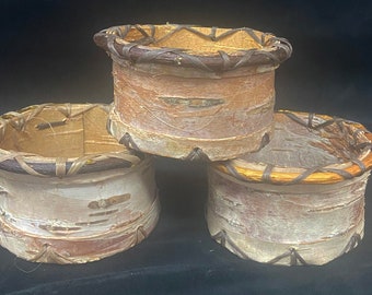 May include: Three round birch bark containers with woven rims. The containers are stacked on top of each other. The top container has a brown rim, the middle container has a brown rim, and the bottom container has a tan rim.