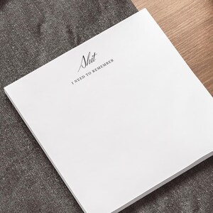 May include: A white square notepad with the text "Shit I NEED TO REMEMBER" in black script. The notepad is on a dark gray surface, with a laptop and a pen visible in the background. A potted plant is also in the background.
