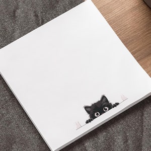 Puede incluir: Un bloc de notas cuadrado blanco con una ilustración de un gato negro asomándose por el borde. El gato tiene ojos grandes y redondos y un aspecto esponjoso. El bloc de notas está sobre una superficie gris oscuro, cerca de un portátil y una planta en maceta.