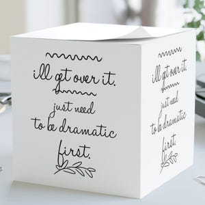 Può includere: Un blocco note bianco a forma di cubo con la scritta "I'll get over it, just need to be dramatic first" in nero. Il blocco note è su una scrivania bianca con graffette e un computer sullo sfondo.