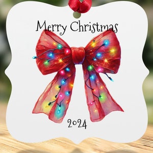 Può includere: Ornamento natalizio bianco con un nastro rosso. L'ornamento presenta un dipinto ad acquerello di un fiocco rosso adornato con luci colorate. Le parole "Merry Christmas" e l'anno "2024" sono stampate.