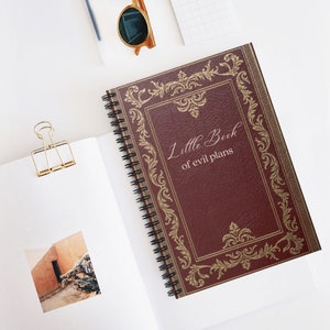 Puede incluir: Una libreta de espiral color burdeos con un marco dorado adornado y el texto "Little Book of evil plans" en la portada.