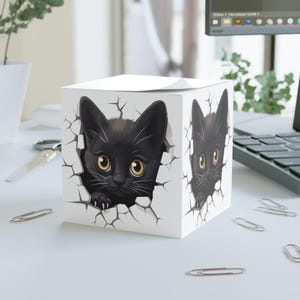 May include: A white cube notepad with a black cat illustration. The cat is depicted breaking through a cracked wall design. The cat has yellow eyes and is looking forward. The notepad is on a white desk with paper clips scattered around.