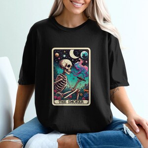 Puede incluir: Camiseta negra con un diseño de carta del tarot. La carta muestra un esqueleto fumando un cigarro, con una colorida ilustración de un cielo nocturno, luna y flores. El texto "THE SMOKER" está en la parte inferior.