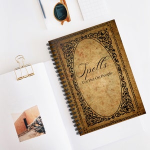 Puede incluir: Un cuaderno de espiral marrón con un diseño vintage. La portada presenta un marco negro adornado con el texto "Spells I've Put On People".