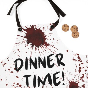 May include: White apron with black ties and the words "DINNER TIME!" in black. The apron features a dark red splatter design. Three chocolate chip cookies are placed next to the apron.