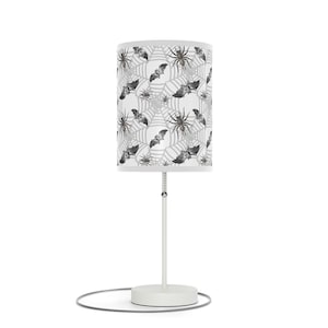 May include: A white table lamp with a cylindrical lampshade featuring a Halloween-themed pattern. The pattern includes grey spiderwebs, spiders, and bats. The lamp has a white base and stem, with a grey cord.