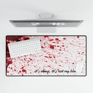 May include: A white keyboard, mouse, and a desk mat with a white background and red splatter design. The desk mat has the text "it's okay, it's not my blood" in a black font. A computer monitor is in the background.