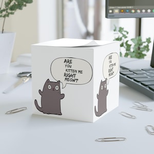 May include: A white cube notepad featuring a cartoon cat and the phrase "ARE YOU KITTEN ME RIGHT MEOW?". The gray cat has big eyes and raised arms. The notepad is on a white surface, with paperclips scattered around.