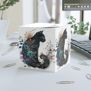 May include: A white cube notepad with a black cat design decorated with colourful floral patterns. The cat is shown in profile, surrounded by teal, purple, and orange flowers and foliage. The design is repeated on the side of the cube.