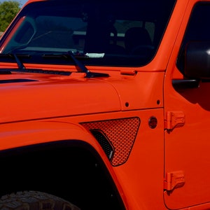 May include: A bright orange Jeep Wrangler with black accents, including a black grille, black side mirrors, and black fender flares. The Jeep has a black windshield wiper and a black door handle.