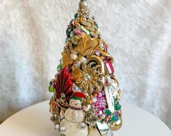 May include: A handcrafted Christmas tree sculpture adorned with an array of vintage jewelry, beads, and ornaments. The tree features a snowman, gold bells, and a pearl-topped spire. The base is covered in glitter, creating a festive holiday decoration.