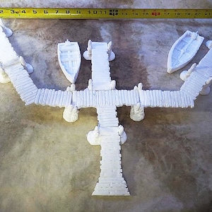 May include: A white miniature pier set with two small boats. The pier is made of white planks and has a Y-shaped design. A yellow measuring tape is placed across the top, showing measurements up to 22 inches.