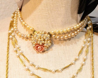 May include: A layered necklace with a pink and white flower charm. The necklace is made of gold chain and pearls.