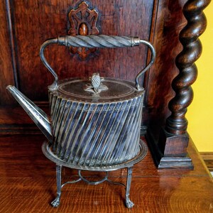 May include: Antique silver-plated tea kettle with a spiral design. The kettle has a decorative handle and a lid with a pineapple-shaped finial. It sits on a matching stand with claw feet, showcasing a vintage aesthetic.