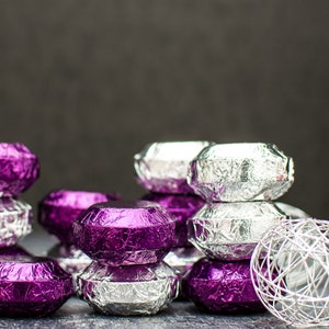 May include: A stack of purple and silver foil-wrapped chocolates. The chocolates are arranged in a pyramid shape, with the silver chocolates on top. There is a white decorative ball in the foreground.