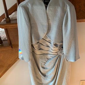 Puede incluir: Conjunto de vestido y chaqueta gris plata claro. El vestido presenta un diseño drapeado con un adorno de joyas en la cintura. La chaqueta es de estilo bolero con mangas largas. El vestido mide aproximadamente 102 cm de largo.