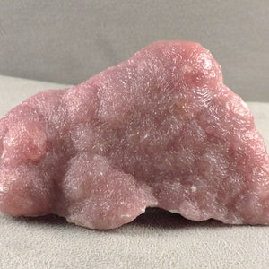 Könnte beinhalten: Ein großer, rosafarbener, botryoidaler Kristallcluster aus rosa Rhodochrosit. Der Kristall hat eine glatte, glänzende Oberfläche und ein durchscheinendes Aussehen.