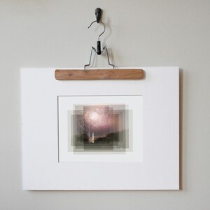 May include: A white framed photograph of a fireworks display over a body of water. The photograph is hanging on a wall with a wooden hanger.
