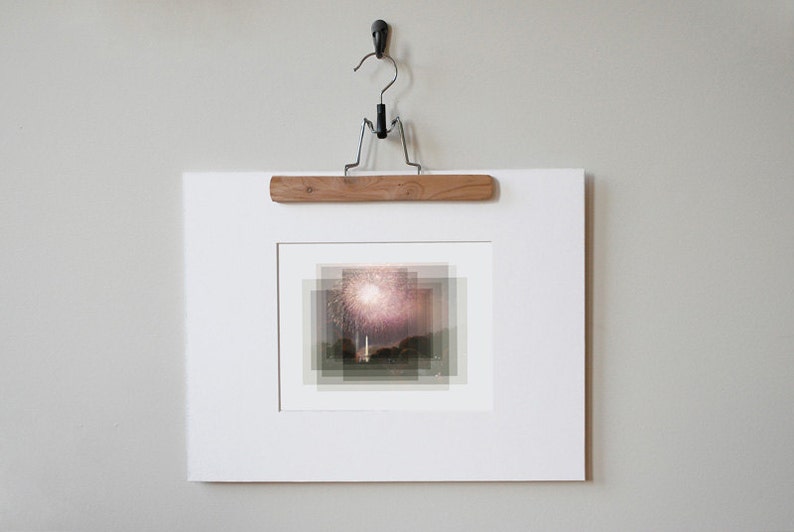 May include: A white framed photograph of a fireworks display over a body of water. The photograph is hanging on a wall with a wooden hanger.