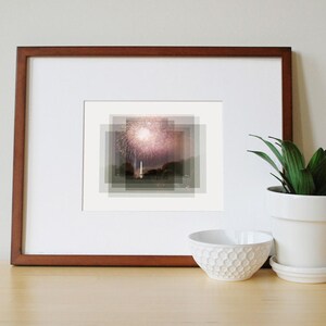 May include: A framed photograph of a fireworks display over a cityscape. The image is in a brown frame and is displayed on a wooden surface.