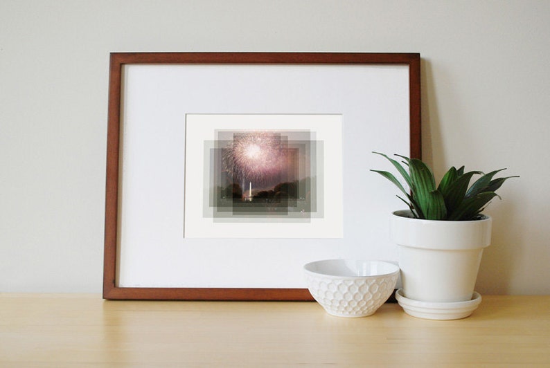 May include: A framed photograph of a fireworks display over a cityscape. The image is in a brown frame and is displayed on a wooden surface.
