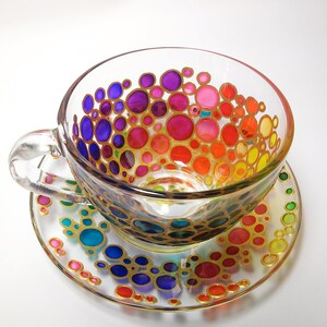 May include: A glass teacup and saucer with a rainbow pattern of colorful circles outlined in gold. The circles are arranged in a gradient from purple to yellow.