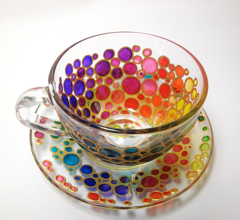May include: A glass teacup and saucer with a rainbow pattern of colorful circles outlined in gold. The circles are arranged in a gradient from purple to yellow.