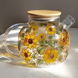 May include: A clear glass teapot with a bamboo lid. The teapot is decorated with hand-painted yellow sunflowers and green leaves. The teapot has a curved handle and a spout for pouring.