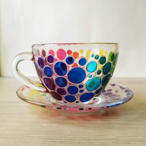May include: A clear glass teacup and saucer with a colorful hand-painted design of rainbow-colored circles.