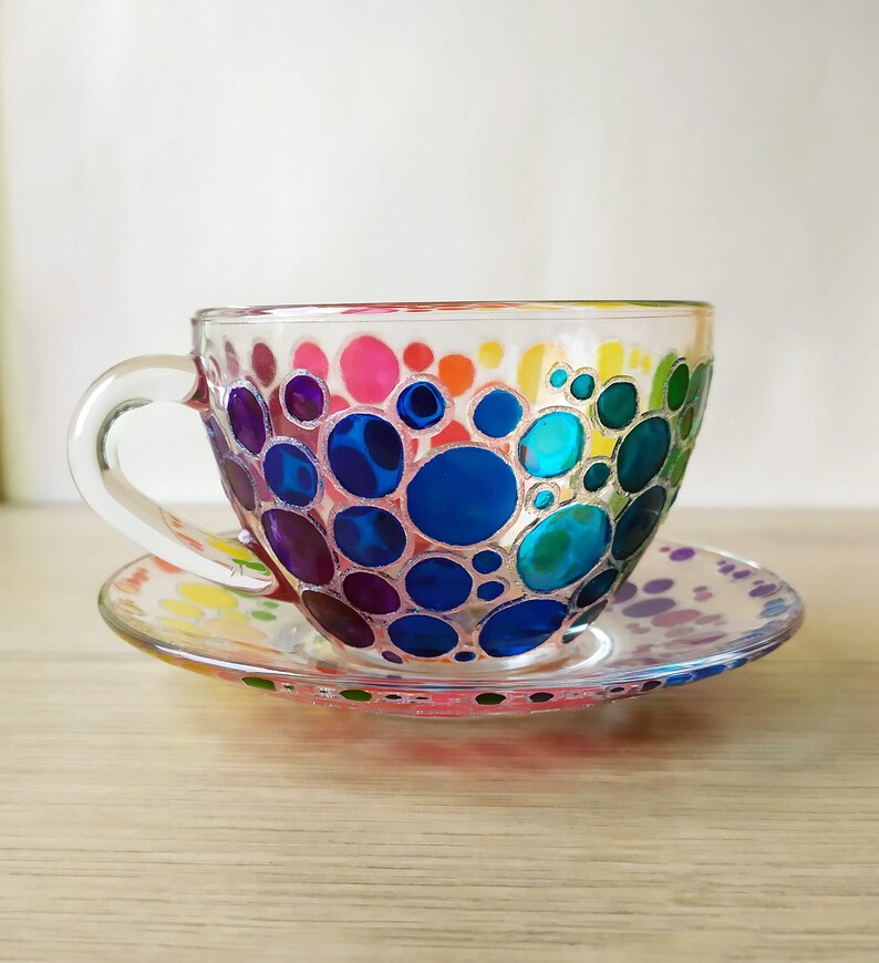 May include: A clear glass teacup and saucer with a colorful hand-painted design of rainbow-colored circles.