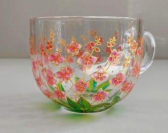 Puede incluir: Una taza de cristal transparente con un diseño pintado a mano de flores de orquídea rosas con contornos dorados y hojas verdes en la base. El patrón floral envuelve el exterior de la taza, que incluye un asa de cristal en el lado derecho.