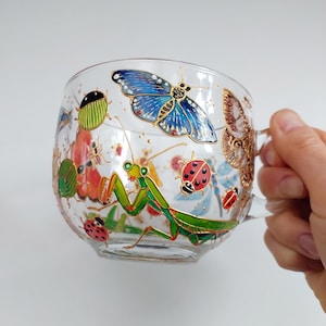 Peut inclure: Une tasse en verre transparent avec une poignée, peinte à la main avec une mante religieuse verte, un papillon bleu, des coccinelles rouges et d'autres insectes. La tasse est décorée d'accents dorés.