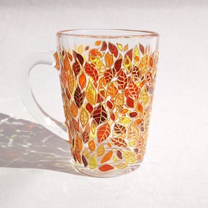Pode incluir: Uma caneca de vidro transparente com alça, decorada com um padrão de folhas de outono pintadas em tons de laranja, amarelo e vermelho.