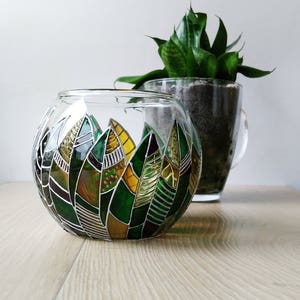 Puede incluir: Un portavelas redondo de vidrio con un diseño de vitral con hojas verdes, amarillas y marrones. El portavelas está sobre una superficie de madera.