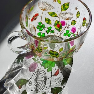 Könnte beinhalten: Eine klare Glas-Tasse mit handgemaltem Design aus grünen Kleeblättern, rosa Blüten und roten Marienkäfern. Die Tasse hat einen goldenen Rand und ist mit weißen Punkten verziert.