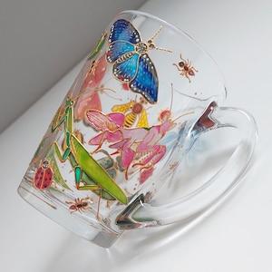 May include: Clear glass mug decorated with colorful insects, including a blue butterfly, green praying mantis, and red ladybug. The mug has a handle and is set against a white background.