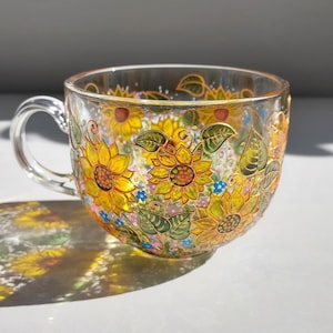 Puede incluir: Una taza de té de vidrio transparente con asa, adornada con girasoles pintados a mano en tonos amarillos, naranjas y verdes. El diseño floral envuelve la taza, acentuado con pequeñas flores azules y hojas verdes. La taza proyecta una sombra.
