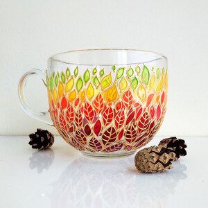 May include: A clear glass mug featuring a hand-painted design of colourful leaves in shades of green, yellow, orange, and red. The leaves are arranged in a gradient pattern, starting with green at the top and transitioning to red at the bottom.
